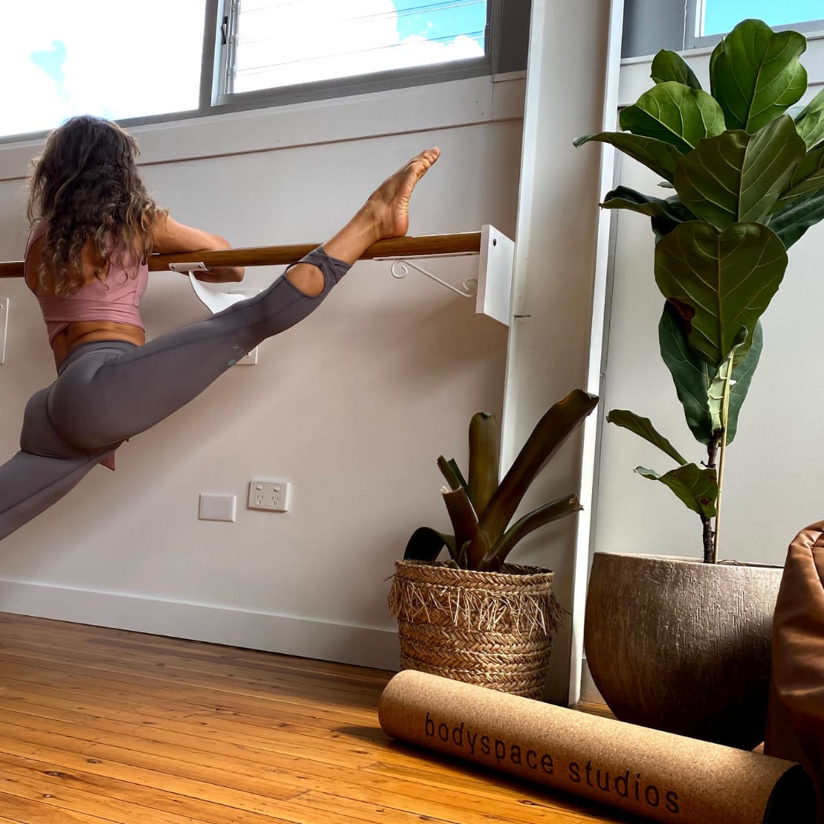 A person performs a split stretch against a barre in a well-lit studio with wooden flooring and indoor plants.