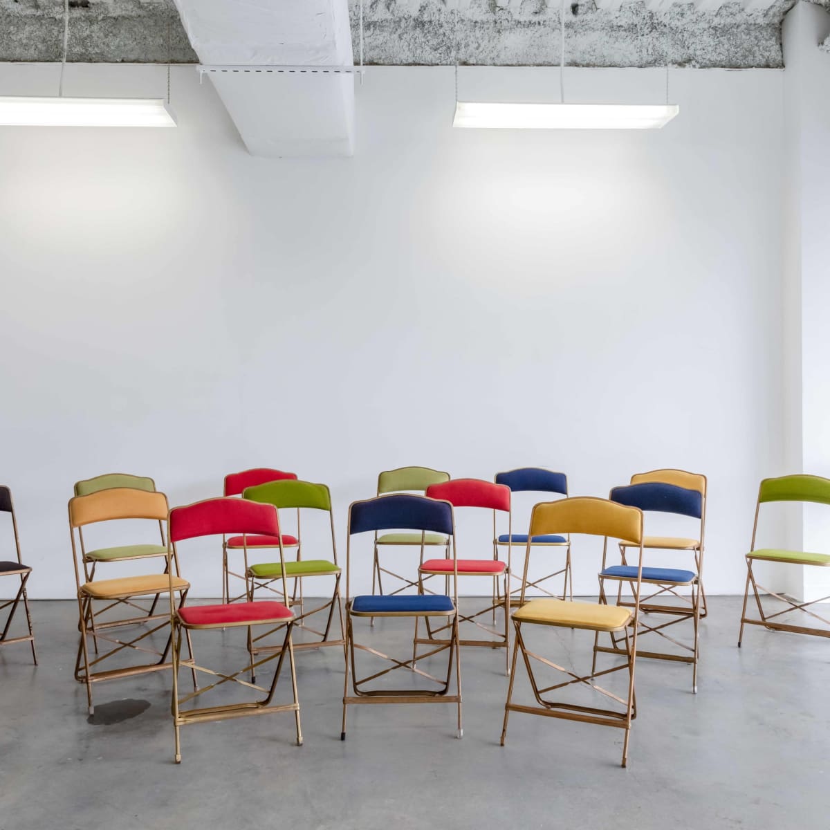 A series of colorful folding chairs are arranged in front of a white wall in a bright, minimalist room.