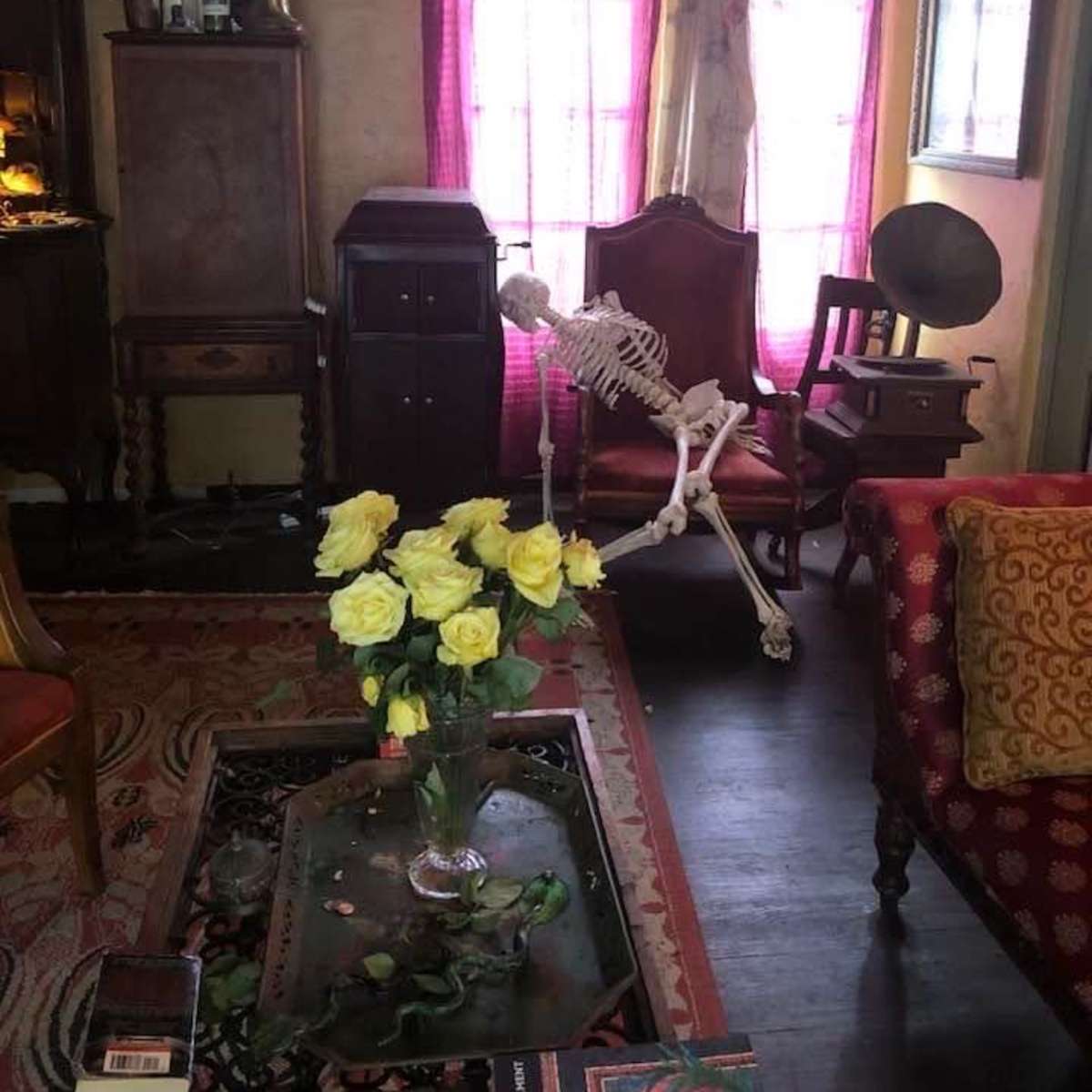 A skeleton sits in an ornate chair next to a vase of yellow roses in a vintage-style room with wooden furniture and colorful curtains.
