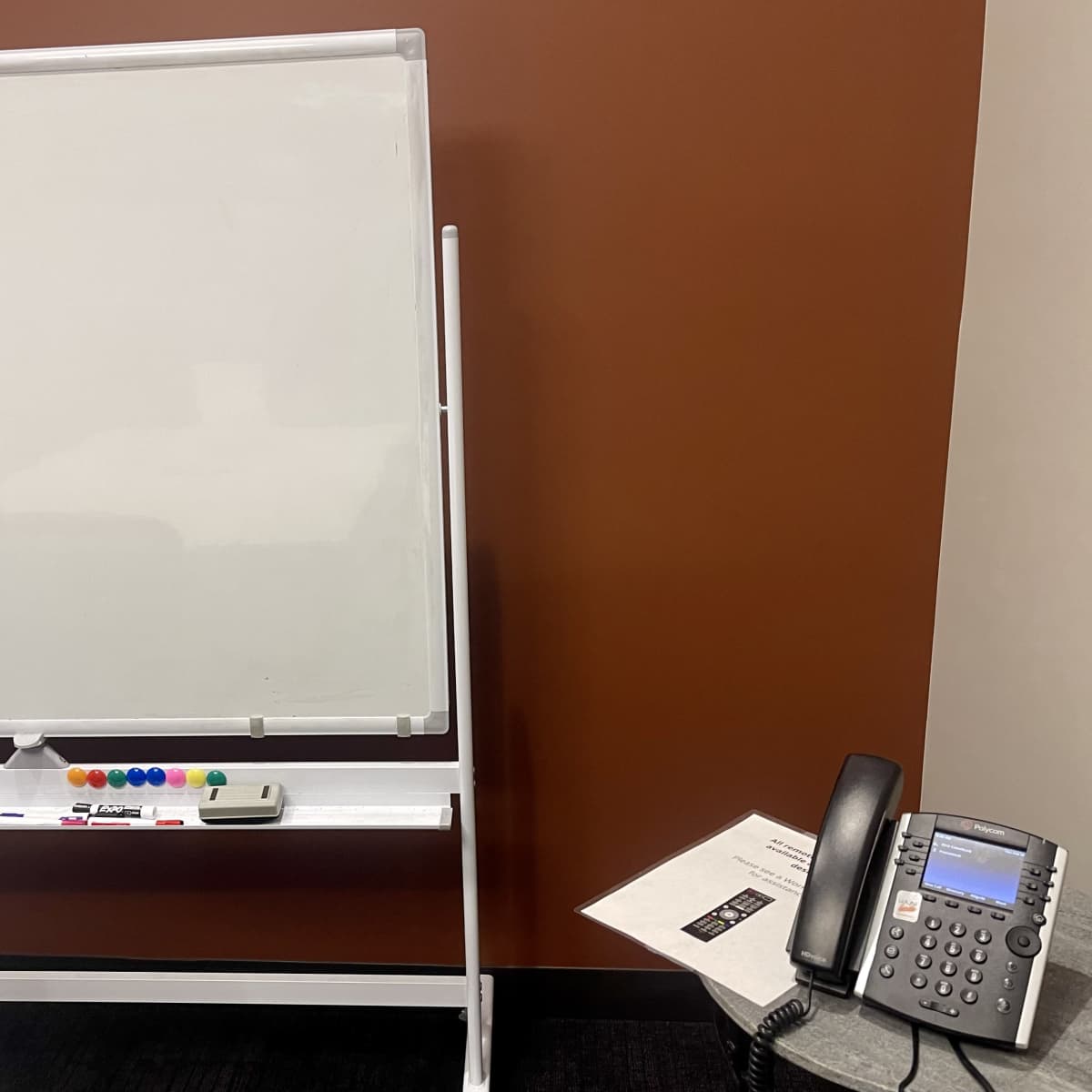 A whiteboard on a stand next to a desk with a telephone and a piece of paper.