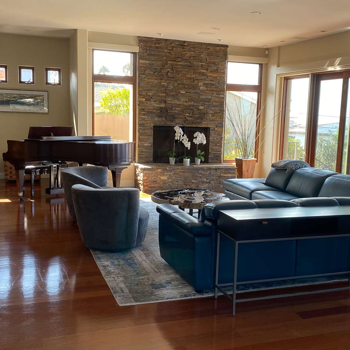 A spacious living room featuring a stone accent wall, a grand piano, a large sectional sofa, and floor-to-ceiling windows that let in ample natural light.
