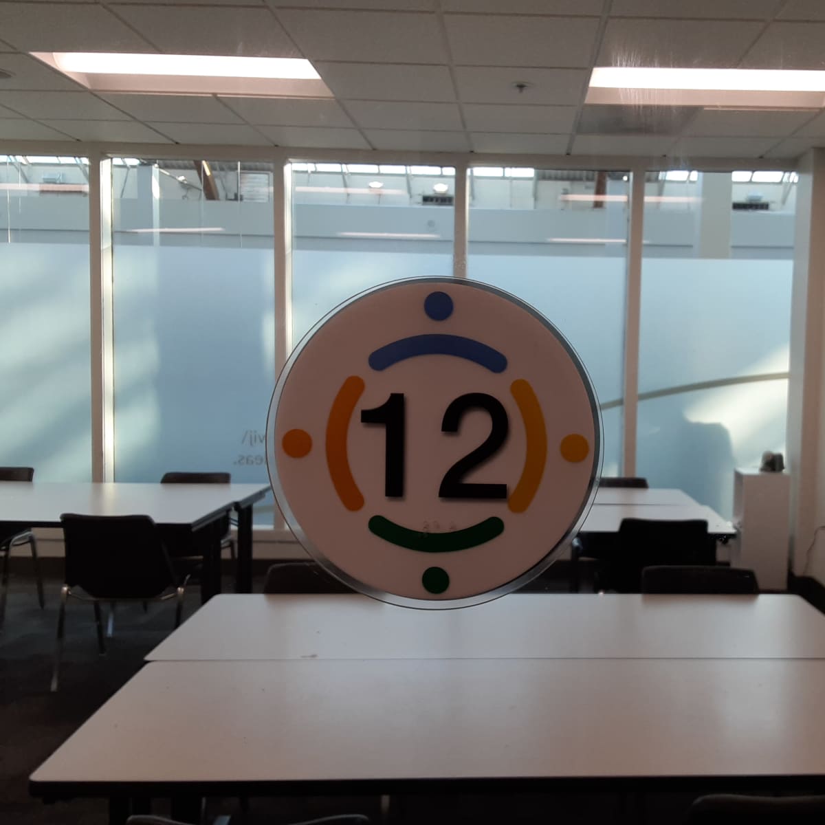 A circular sign with the number "12" in the center, affixed to a glass door leading into a room with several tables and chairs.