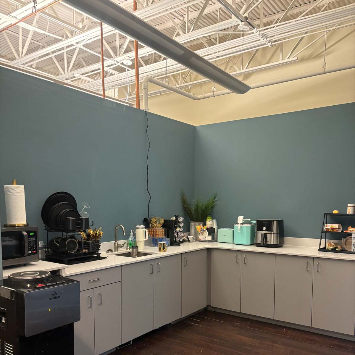 A modern kitchen area with gray cabinets, a coffee station, and various appliances against a blue wall.