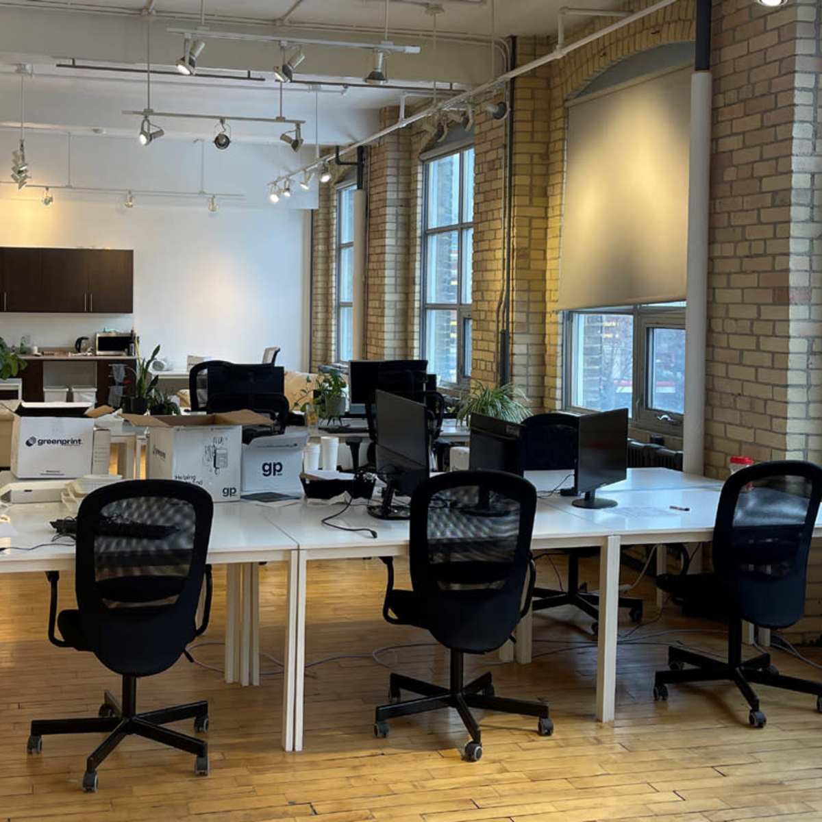 A spacious office with several desks, computer chairs, and large windows, featuring a lounge area in the background.