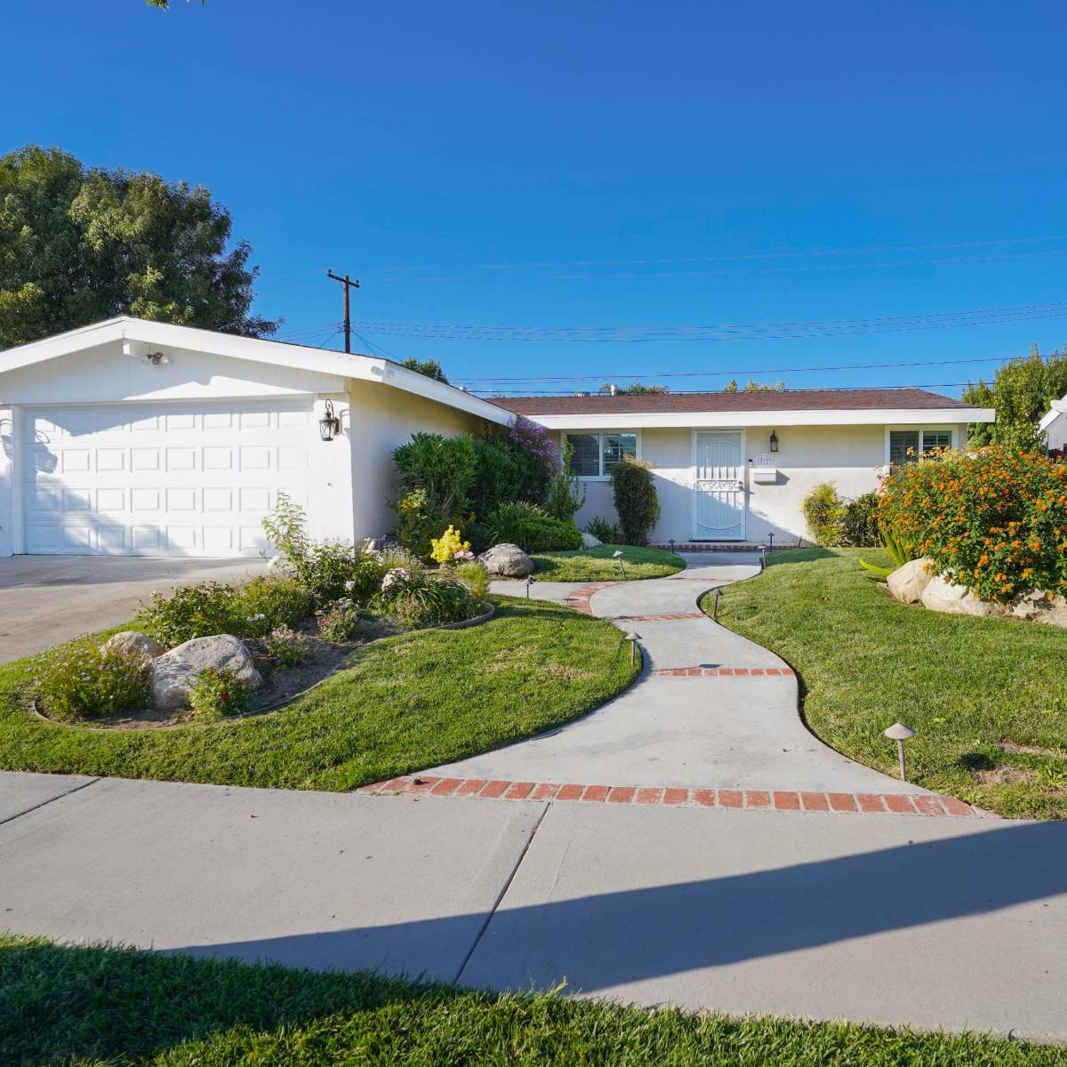 A single-story house with a garage is surrounded by a landscaped yard featuring a curved pathway and various plants.