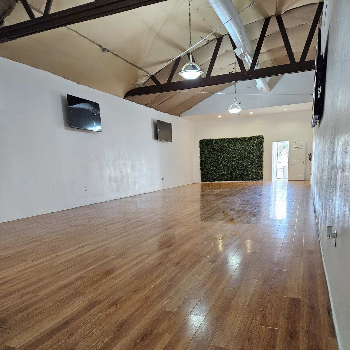 A spacious, empty room with hardwood floors, equipped with two televisions mounted on the walls and a green plant wall feature at one end.