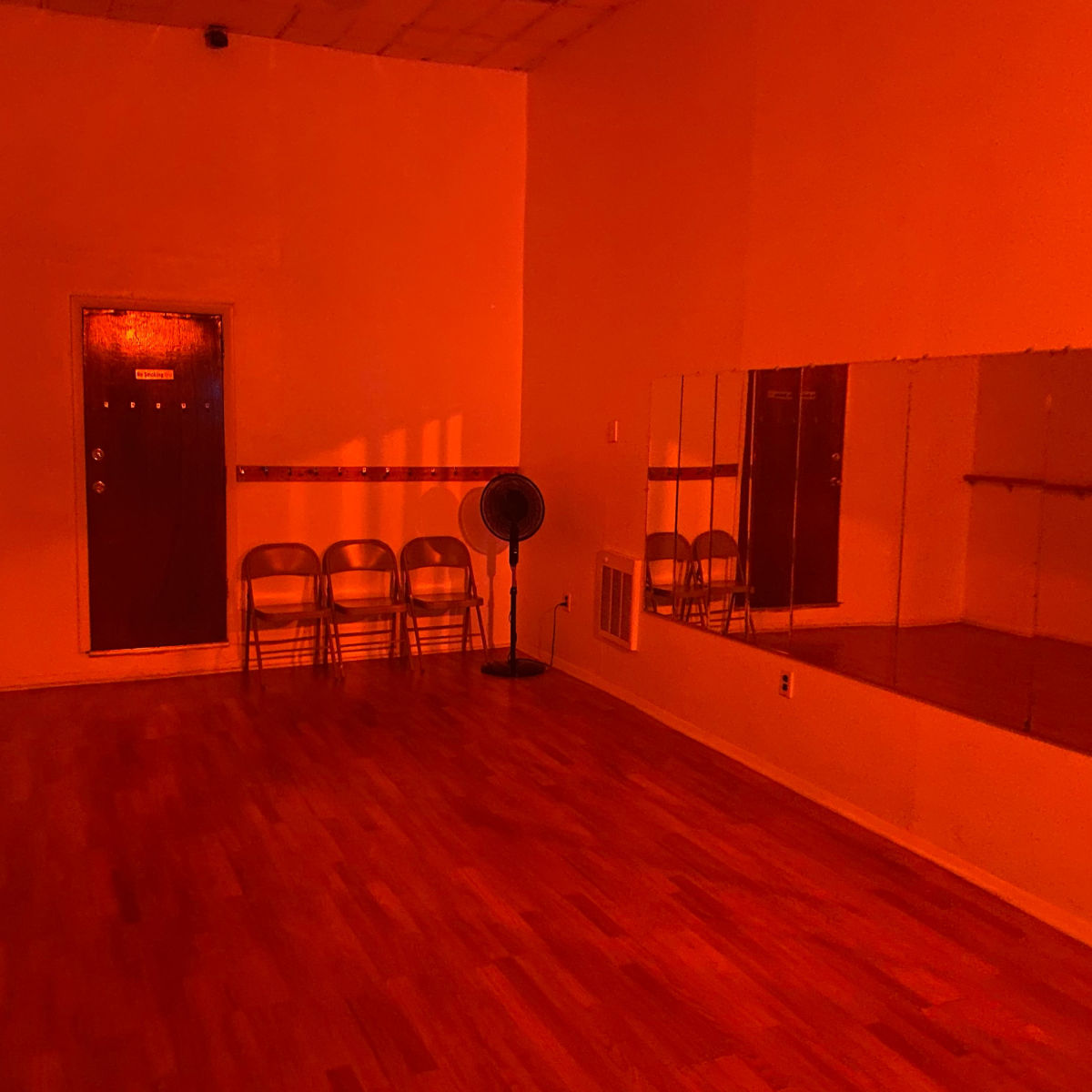 An empty room with wooden flooring, four metal chairs lined up against the wall, a standing fan, and a mirrored wall, all illuminated by a reddish light.