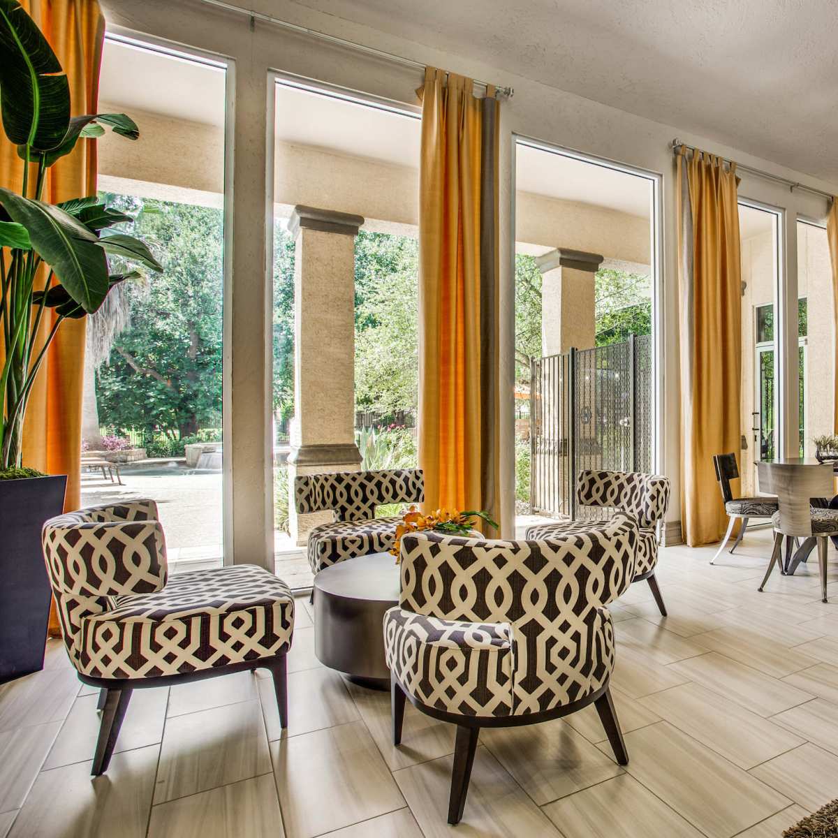 A spacious lounge area with patterned chairs, a round table, and workstations, surrounded by large windows and bright curtains.