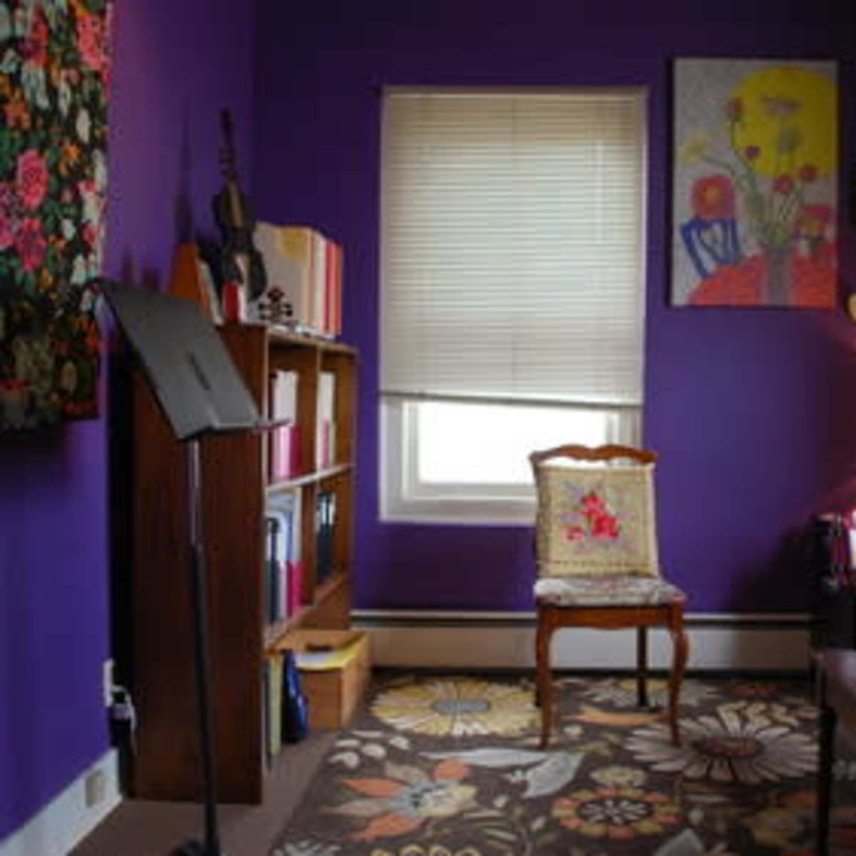 A small room with purple walls, a chair, a bookshelf, and a piano, featuring a floral rug and various artworks on the walls.