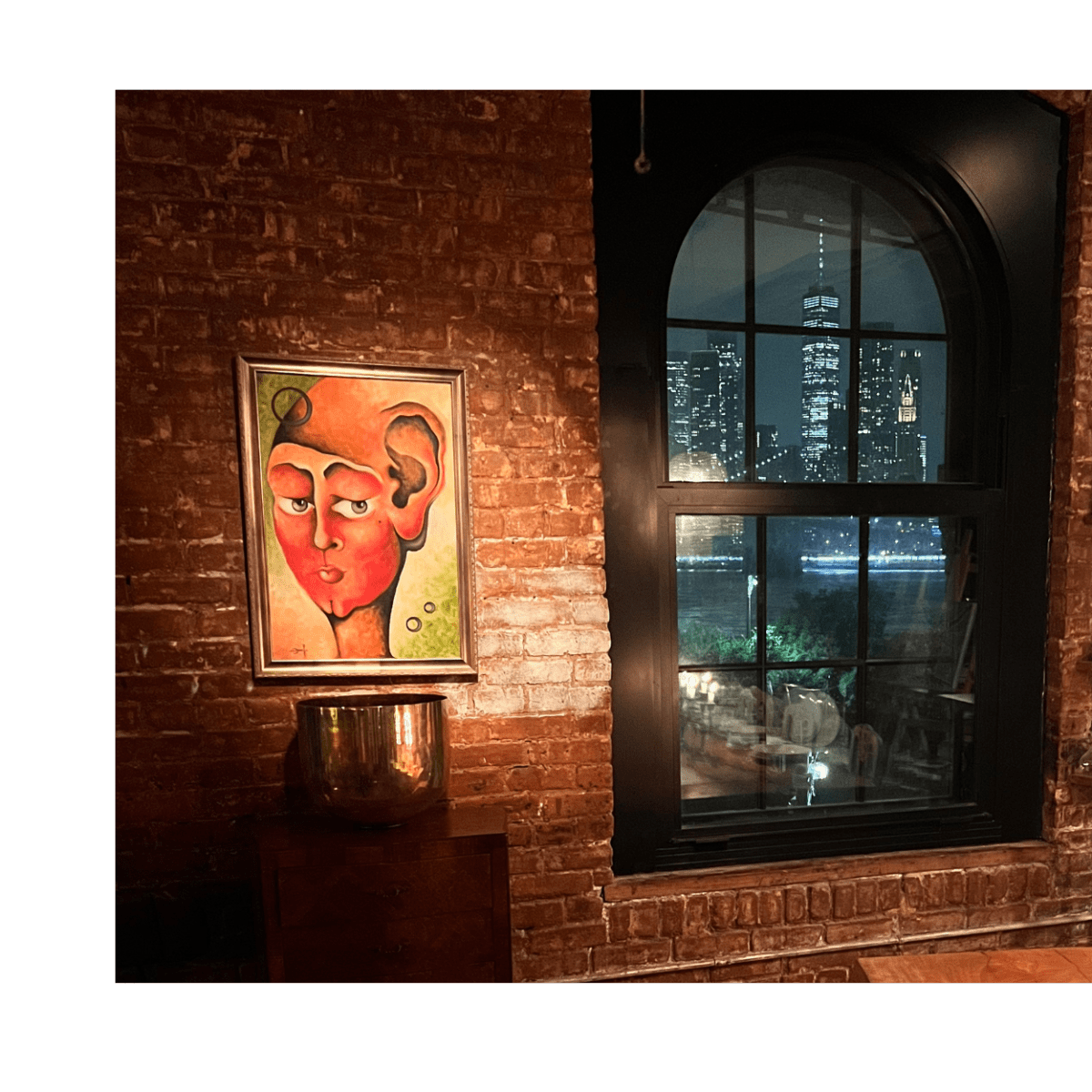 A brick-walled interior with a framed painting of a face, illuminated by warm lighting from hanging fixtures, overlooking a city skyline through a large arched window.