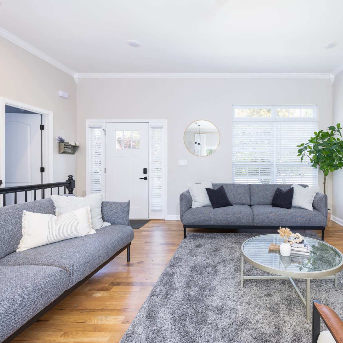 A bright living room features two gray sofas, a glass coffee table, and a large wall-mounted television.