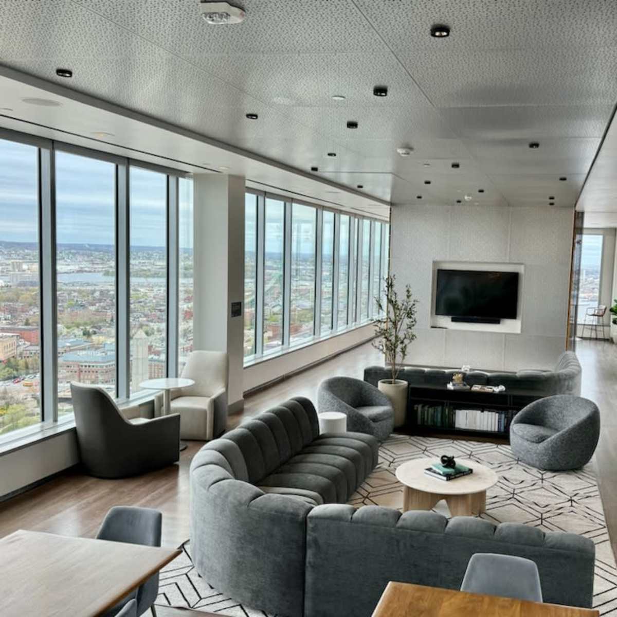 A modern lounge with large windows offering a city view, featuring gray furniture and a central coffee table.