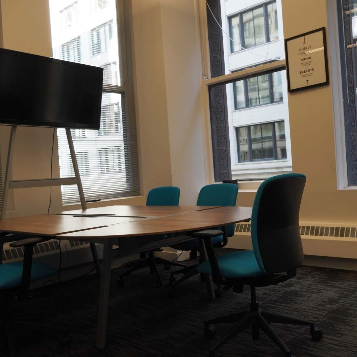 A meeting room features a wooden table surrounded by teal chairs and a mounted television, with large windows allowing natural light.