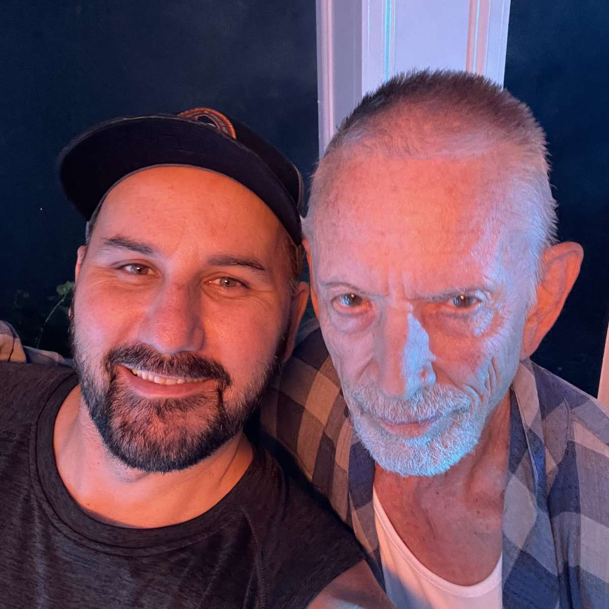 Two men pose together for a selfie, with one wearing a baseball cap and the other in a plaid shirt.