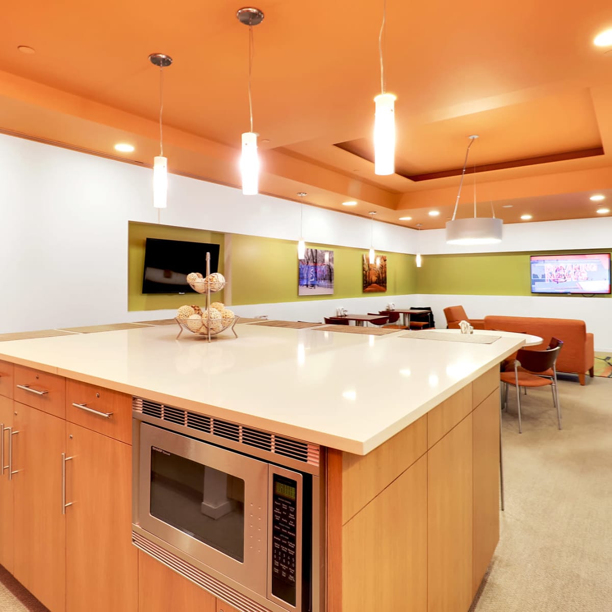 A modern kitchen features an island with a microwave and a seating area, complemented by orange and green walls and recessed lighting.