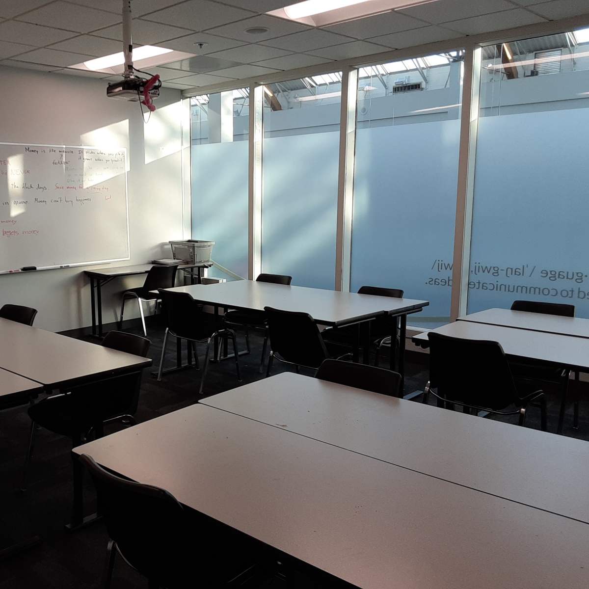 A classroom with several tables and chairs arranged around a whiteboard and large windows allowing natural light to enter.