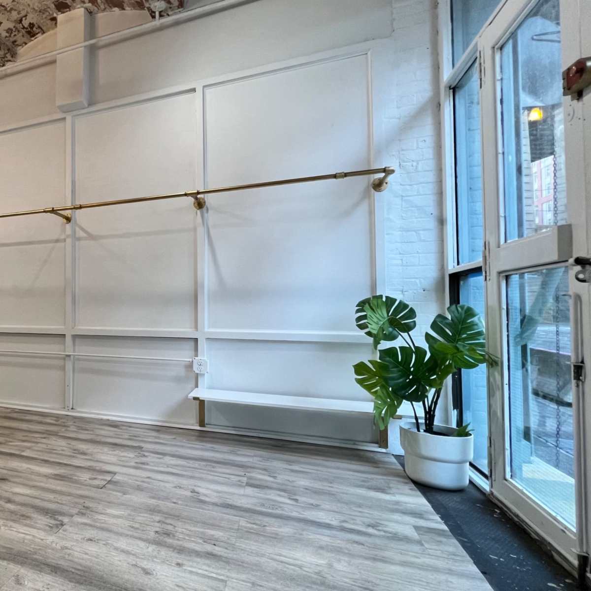 An empty room with a wooden floor, white walls, and a brass clothing rack along one side, illuminated by natural light from large windows.
