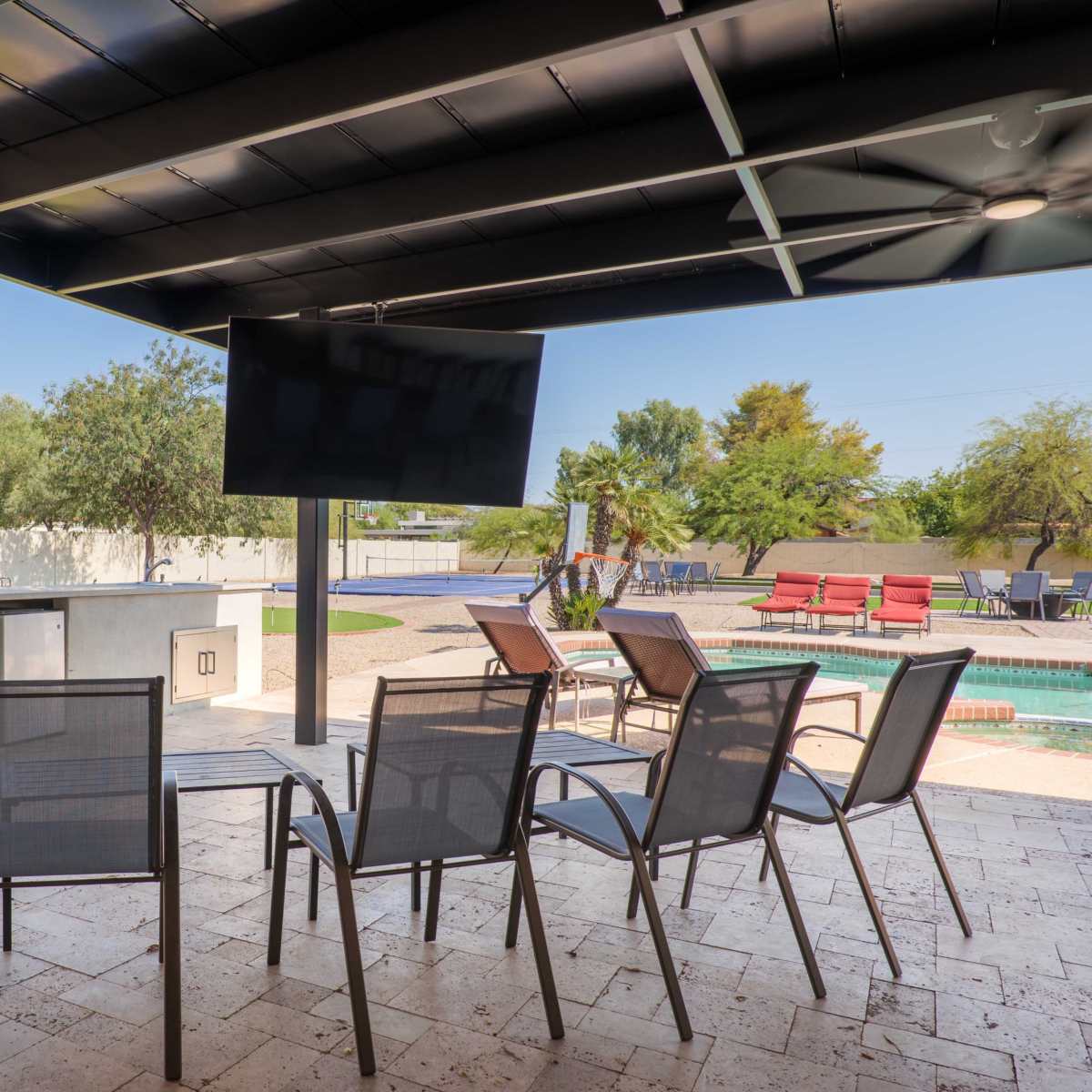 A covered outdoor patio with a barbecue grill, a large television mounted on the wall, and several chairs arranged around a patio table, overlooking a swimming pool and landscaped yard.