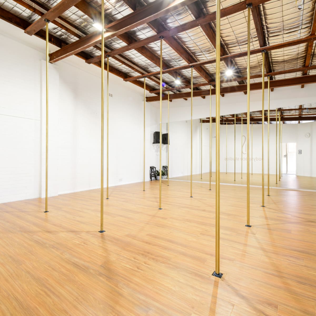 An empty dance studio with wooden flooring and multiple gold poles spaced evenly throughout the room.