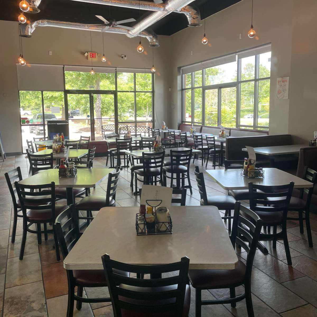 An empty restaurant interior with multiple tables and chairs arranged around a central area, large windows letting in natural light, and industrial-style lighting fixtures.