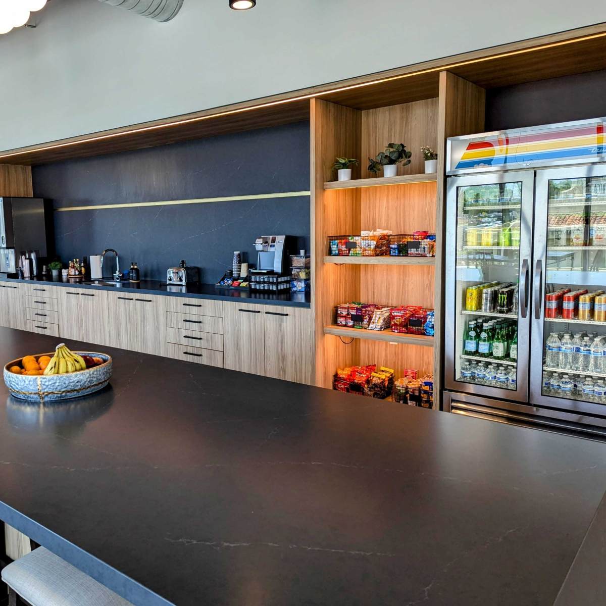 A modern kitchen area featuring a large dark countertop, cabinets, a refrigerator, and a display of snacks and beverages.