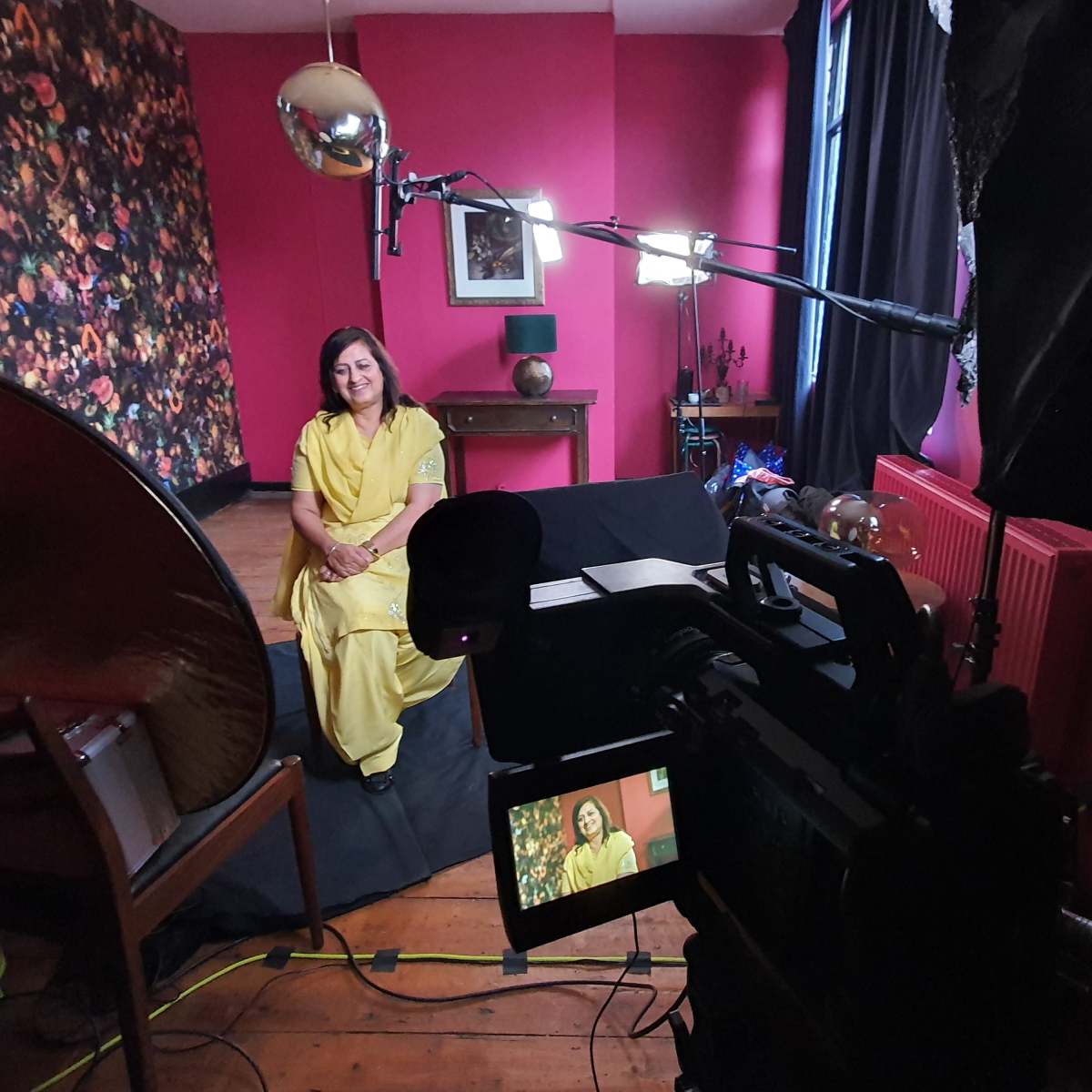 A woman in a yellow outfit sits on a black couch in a room with pink walls and floral wallpaper, while a camera and lighting equipment are set up for recording.