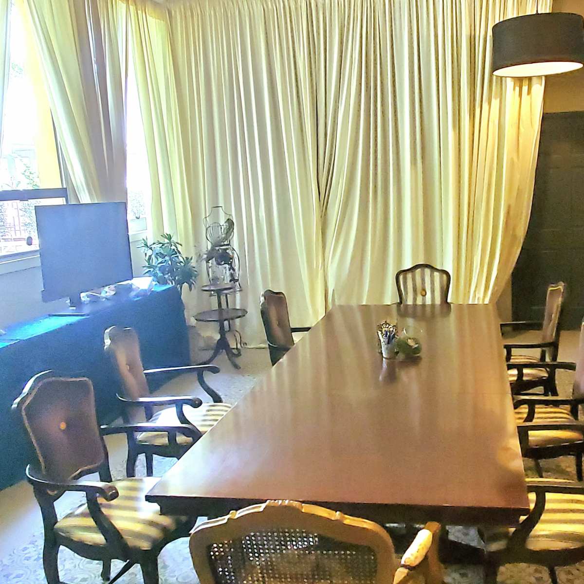 A conference room with a long wooden table surrounded by eight chairs, large windows covered with yellow curtains, and a doorway leading to another room.
