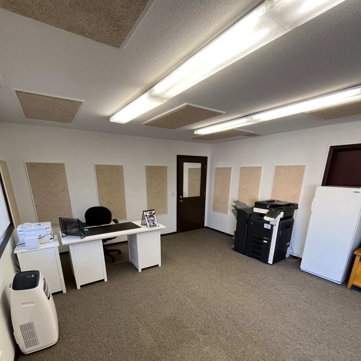 An office space featuring a desk with a chair, a printer, a refrigerator, and carpeted flooring, with soundproof wall panels.