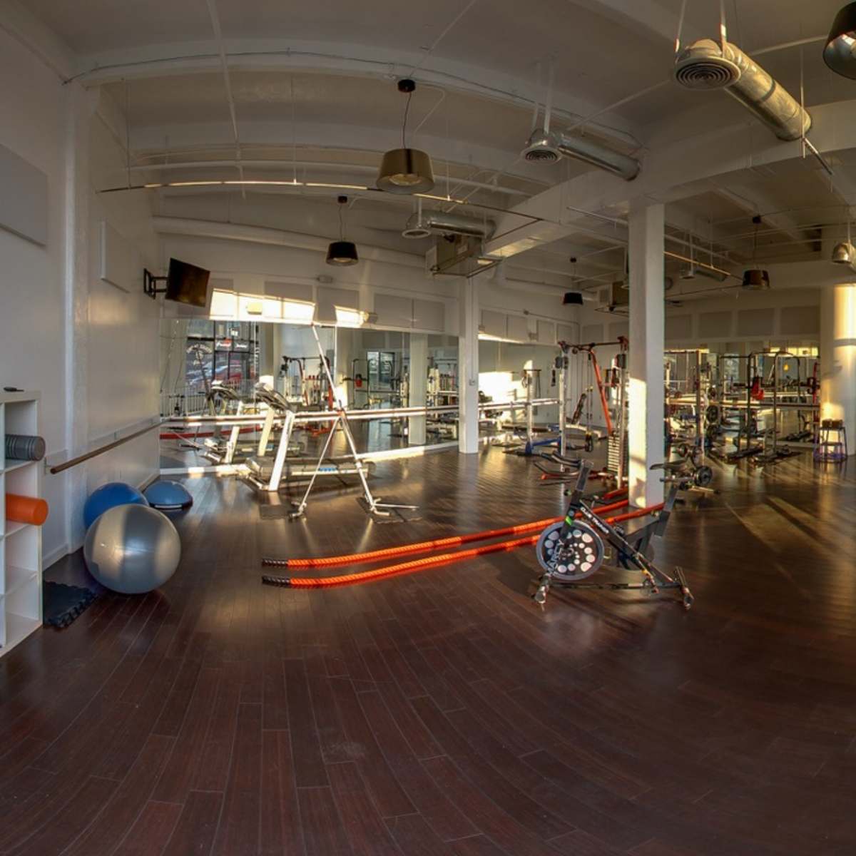A spacious gym interior featuring exercise equipment, mats, and a wooden floor, with natural light coming through large windows.