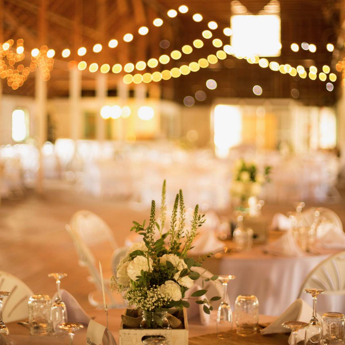 A rustic event space features neatly arranged tables with white linens, a centerpiece of flowers in a wooden box, and string lights illuminating the ceiling.