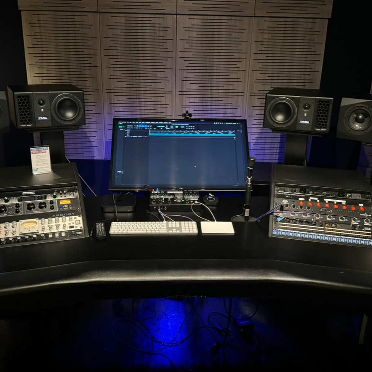 A music production studio setup features a central monitor, audio interface, and studio monitors on either side, with various equipment arranged on a curved desk.