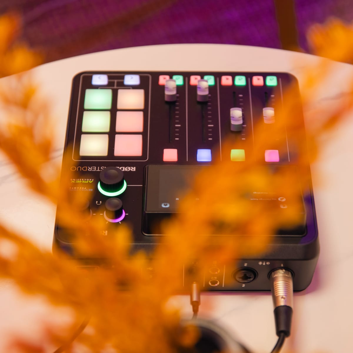 A black audio mixing device with colorful buttons and sliders is positioned on a white table, partially obscured by yellow foliage.