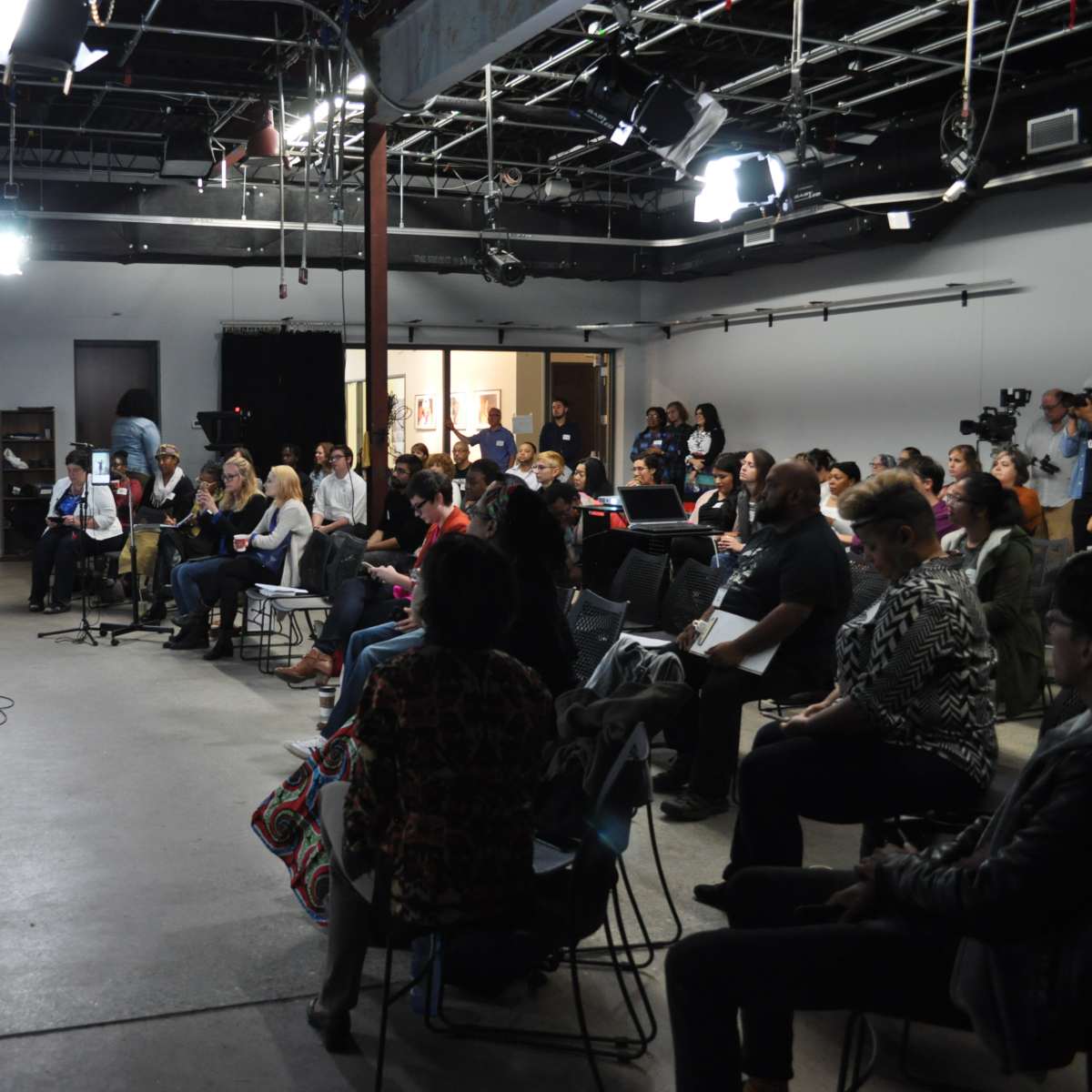 A speaker stands on a stage in front of an audience seated in a modern, industrial-style venue.