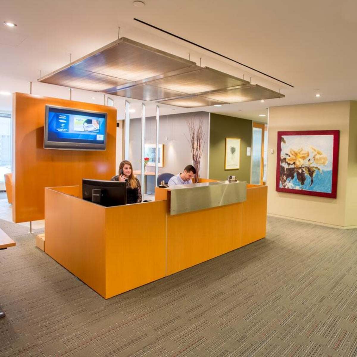 A modern office reception area with two employees at a front desk and a hallway leading to other rooms.