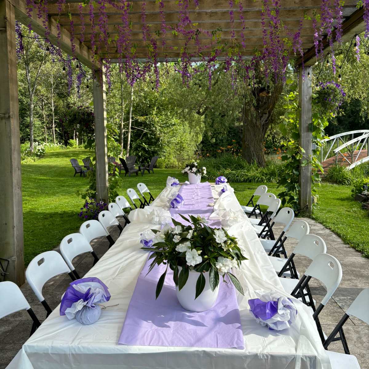 A long table is set under a pergola adorned with hanging purple flowers, surrounded by greenery and white folding chairs.