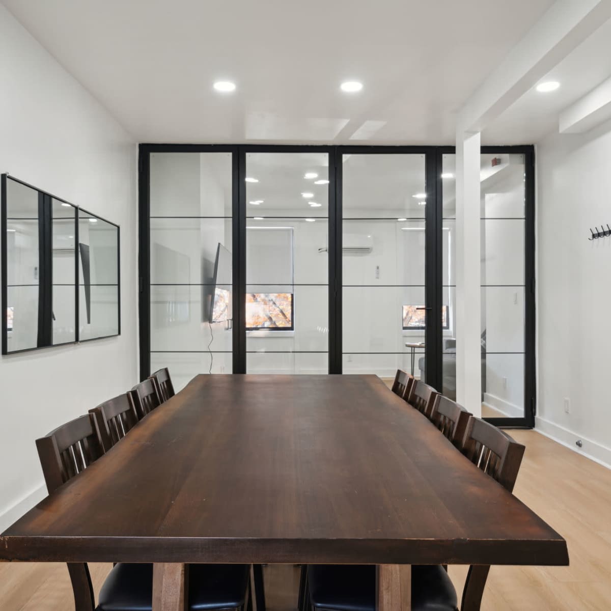 A long wooden dining table with eight chairs is set in a bright room featuring glass walls and a large mirror.