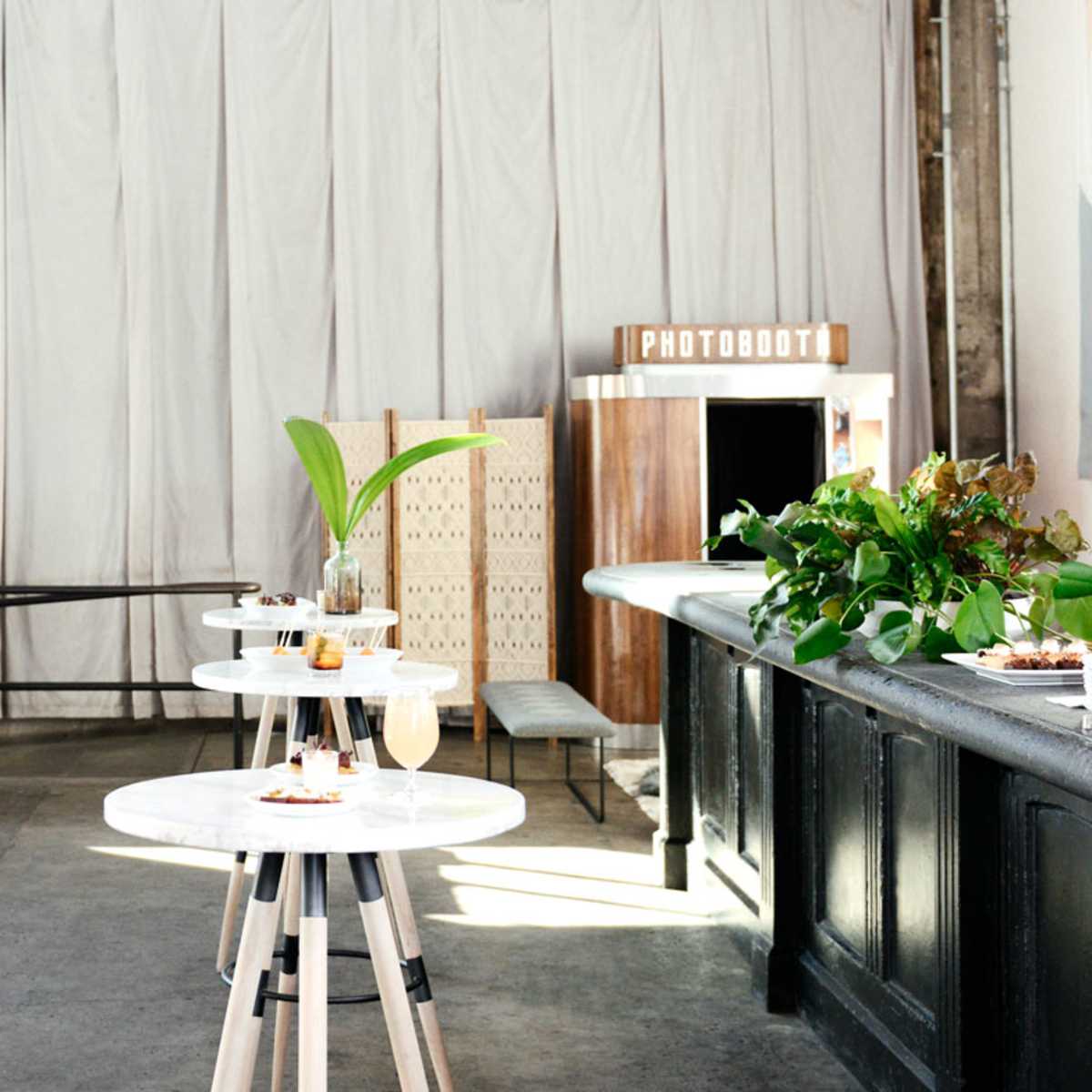A spacious interior with modern furniture, including tables and a couch, alongside a photobooth and a greenery arrangement.