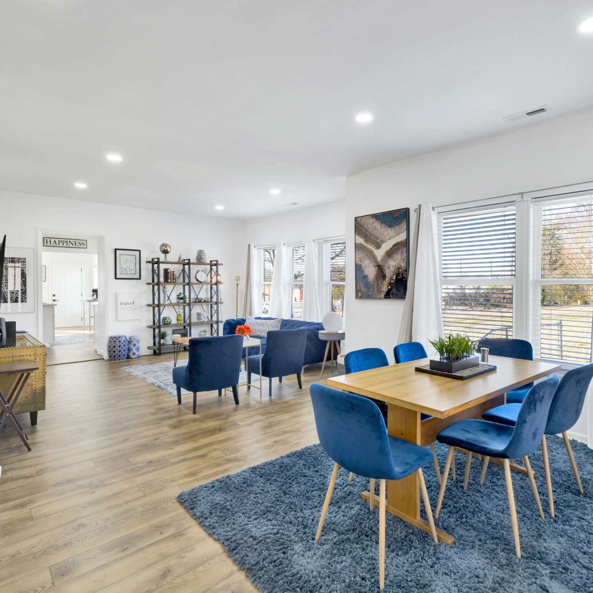 A bright, modern dining area features a wooden table surrounded by blue chairs, with large windows allowing natural light and a view of the outdoors.