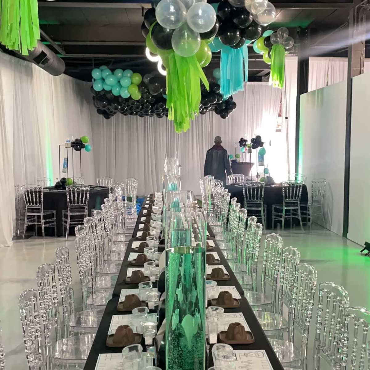 A long banquet table is set with clear chairs and decorative centerpieces, while colorful balloons hang above in an elegantly arranged event space.