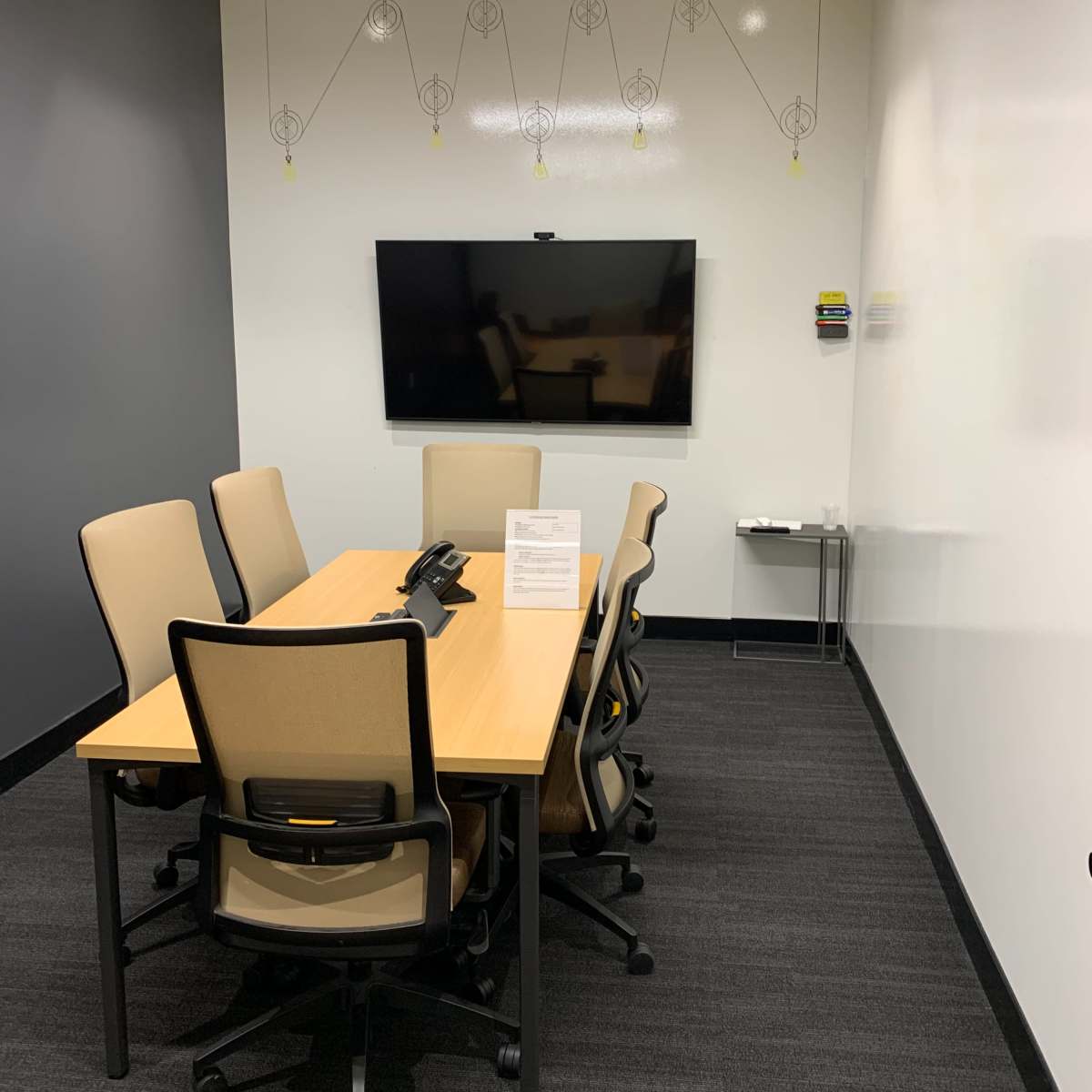 A small conference room with a rectangular table surrounded by six chairs, a wall-mounted television, and a small side table.