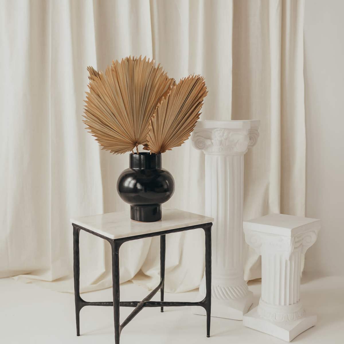 A black vase with dried palm fronds stands on a marble-topped table beside two white columns against a backdrop of cream-colored curtains.