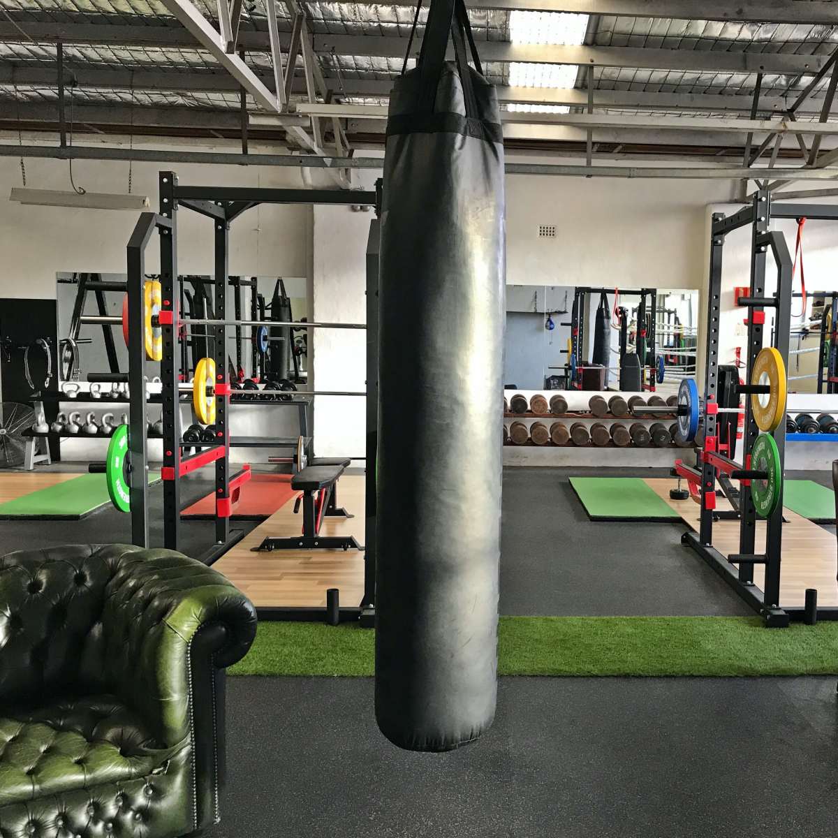 A gym interior featuring weightlifting equipment, a punching bag, and two distinct seating areas with a leather couch and an armchair.