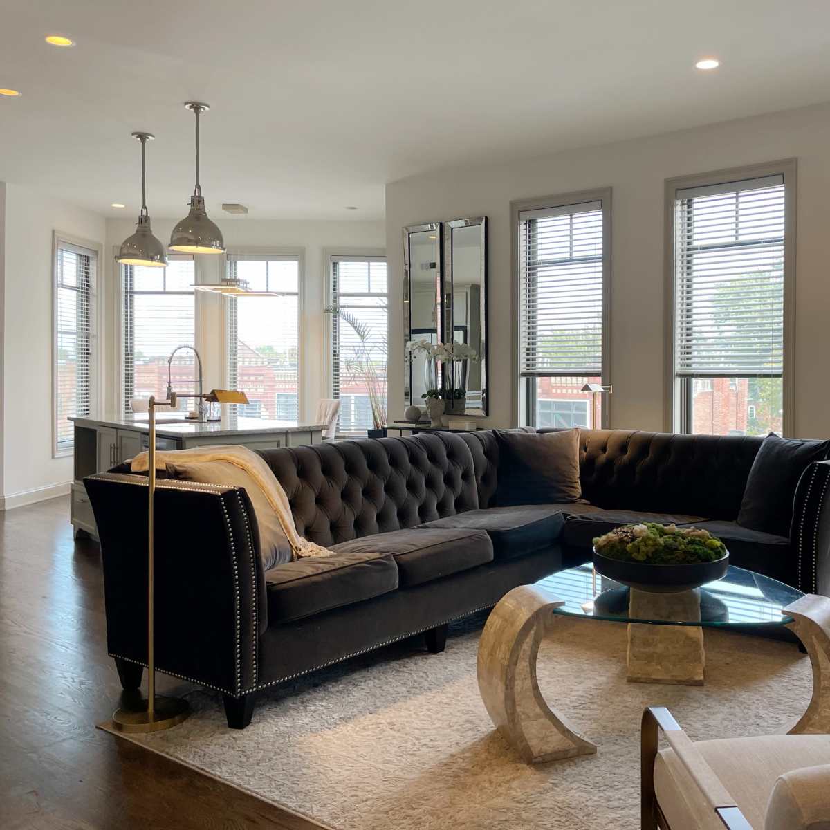 A modern living room with a large tufted sectional sofa, glass coffee table, and bright natural light streaming through multiple windows.