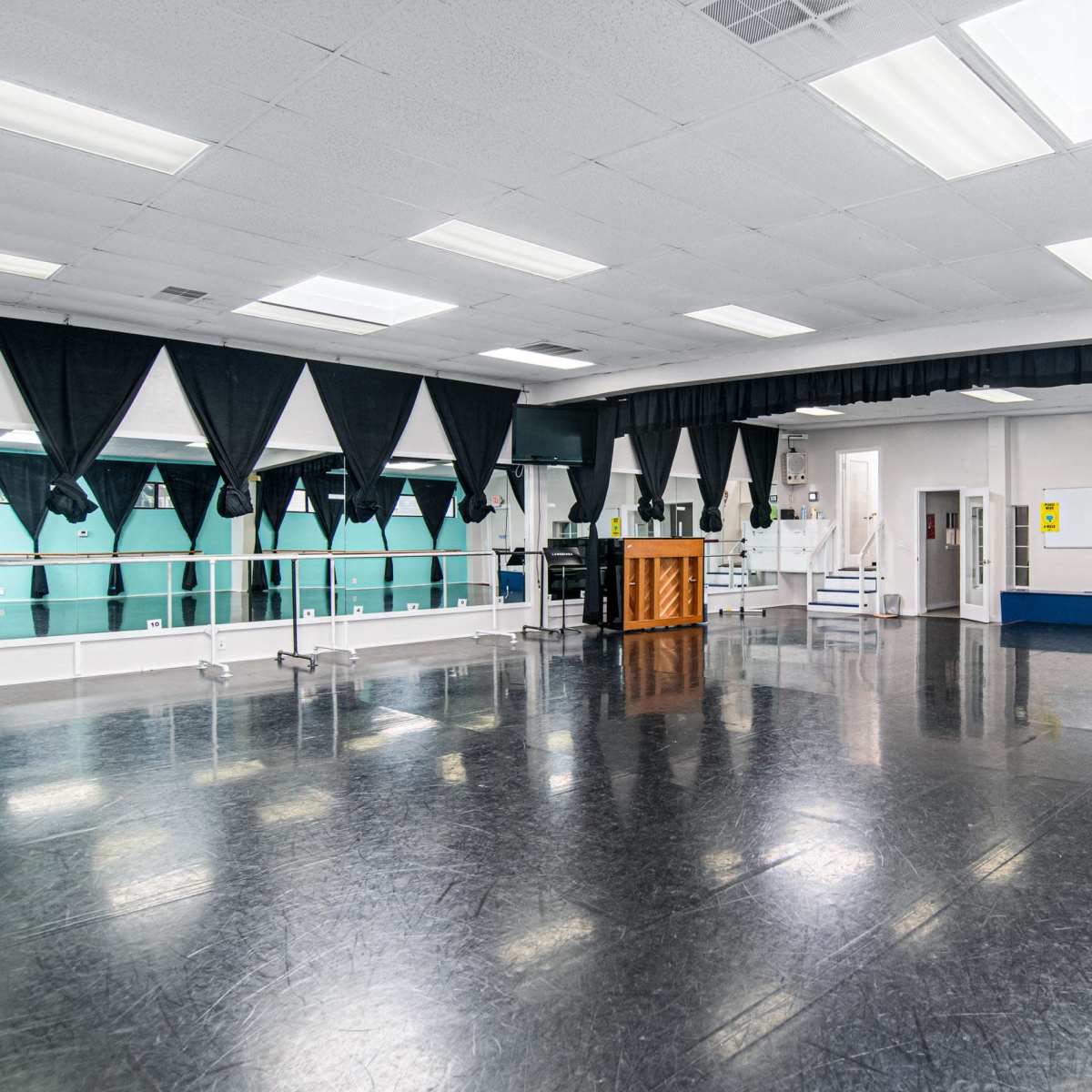 A spacious dance studio with a black floor, large mirrors, and a grand piano, surrounded by curtain-draped windows.