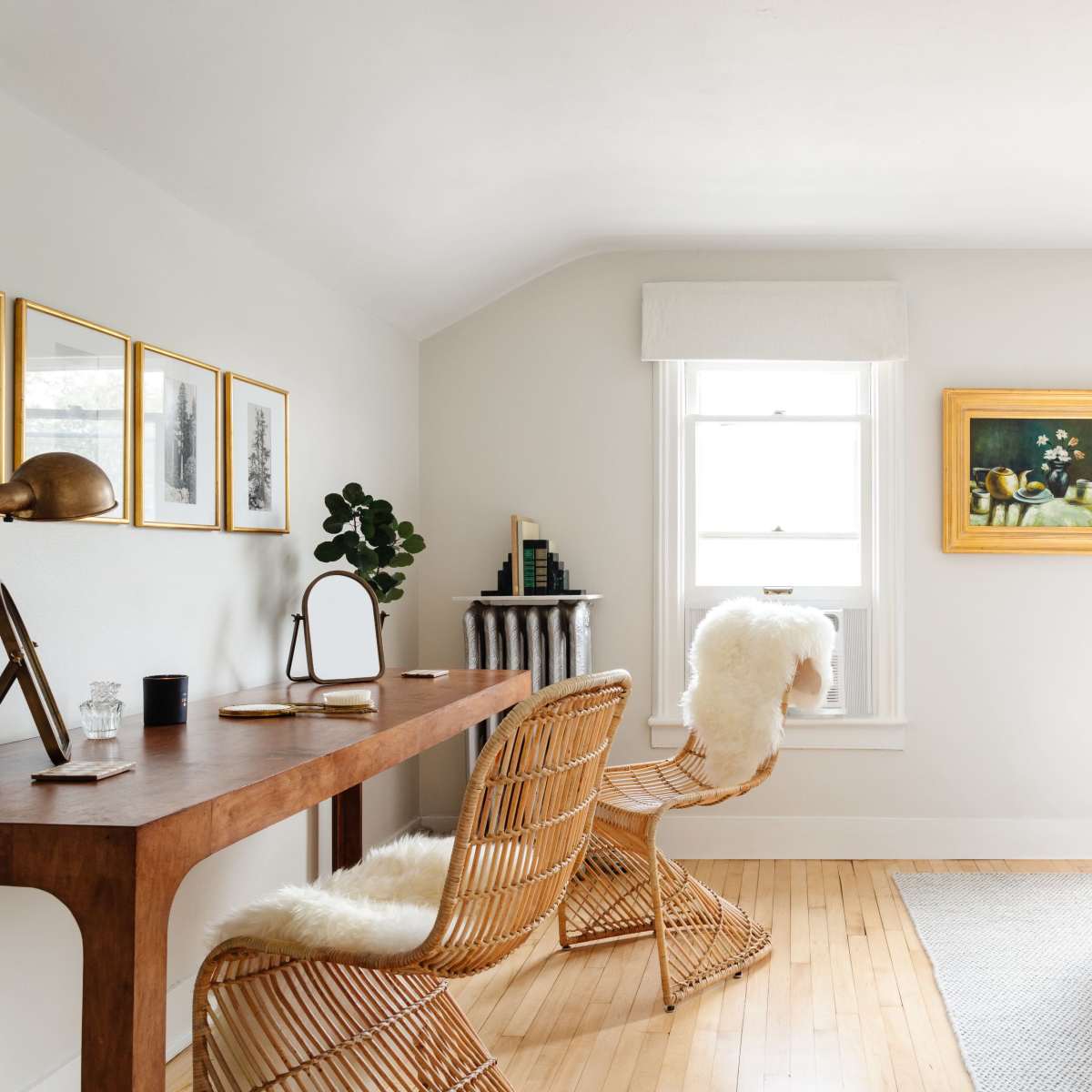 A well-lit bedroom features a wooden desk with a chair, a bed with a light quilt, and artwork displayed on the walls.