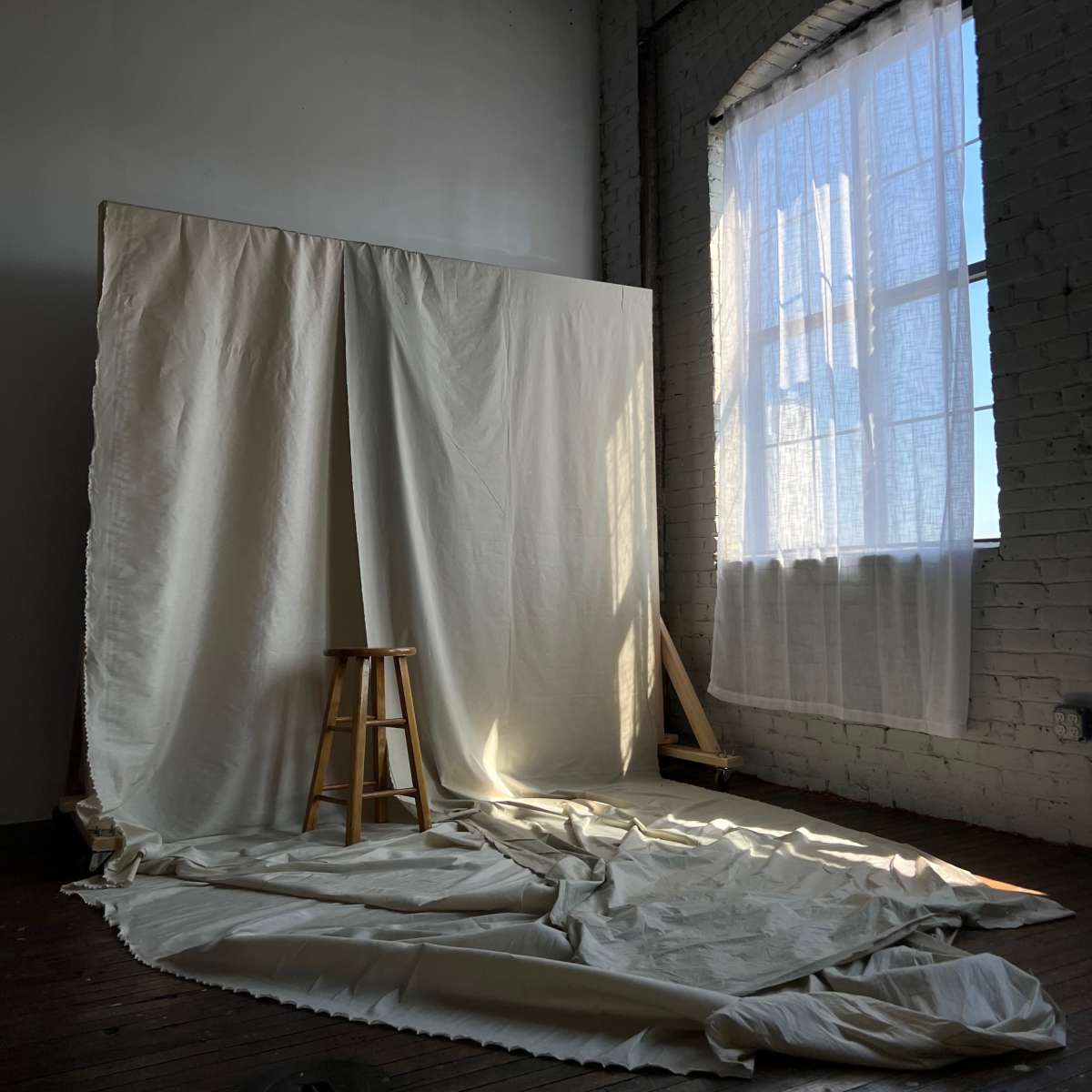 A simple room features a large white fabric backdrop and a wooden stool beside a window with sunlight streaming through.