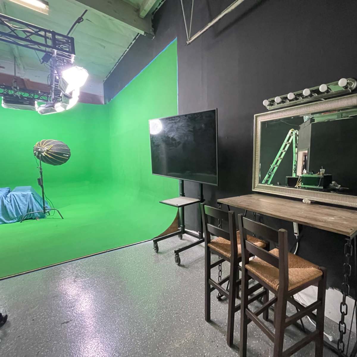 A studio space featuring a green screen backdrop, a makeup mirror with lights, a small table with two chairs, and a television on a stand.