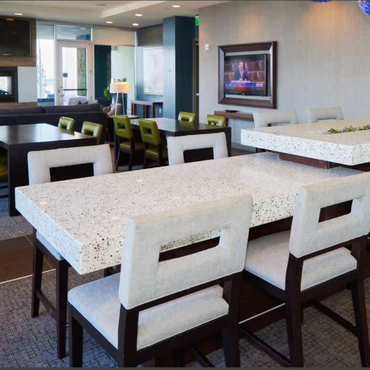 A modern dining area with a large white terrazzo table surrounded by upholstered chairs, with additional seating and large windows in the background.