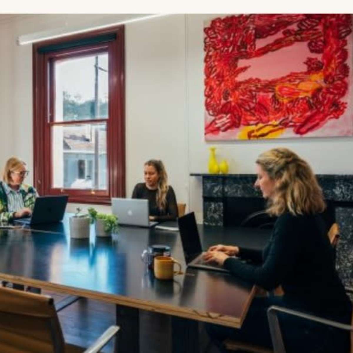 Three people work on laptops at a large conference table in a brightly lit meeting room with large windows and modern decor.