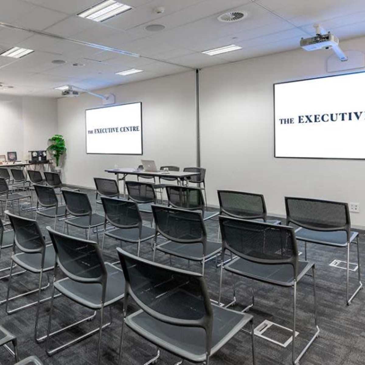 A modern conference room with rows of black chairs, a projection screen displaying "THE EXECUTIVE CENTRE," and a refreshment table along one wall.
